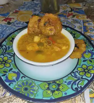 Bowl of savory festival stew with shrimp, vegetables, potatoes, and fried plantain sides.